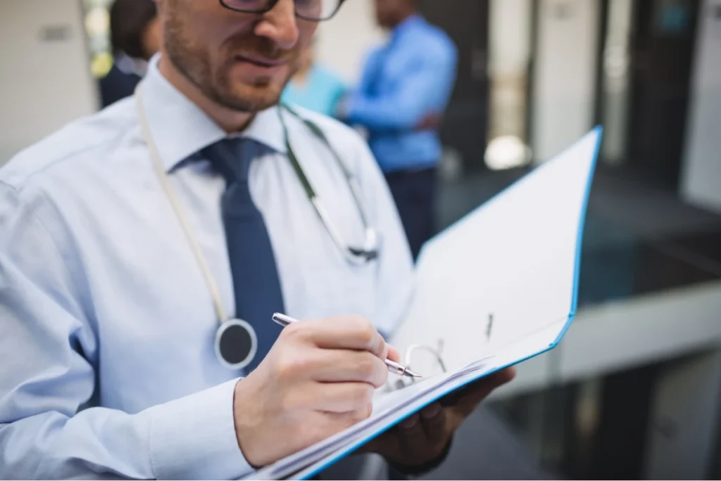Doctor writing notes on a clipboard, wearing a stethoscope around the neck.