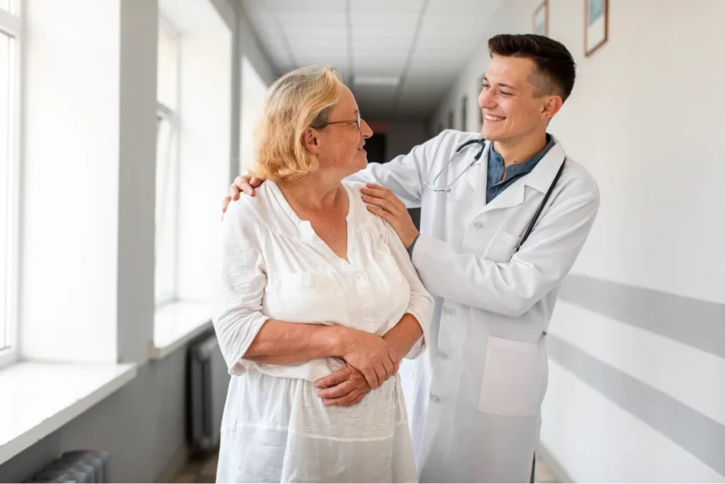 Smiling doctor with a stethoscope comforting an elderly woman in a hospital hallway.