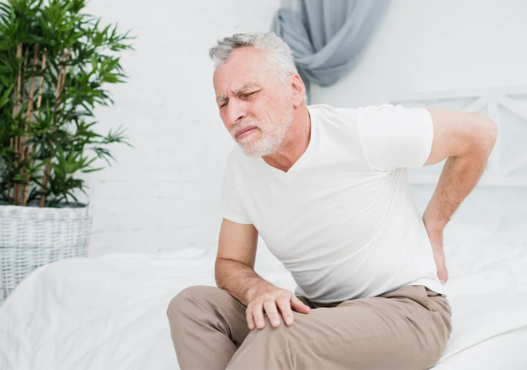 Older man sitting on bed, holding his lower back in pain.