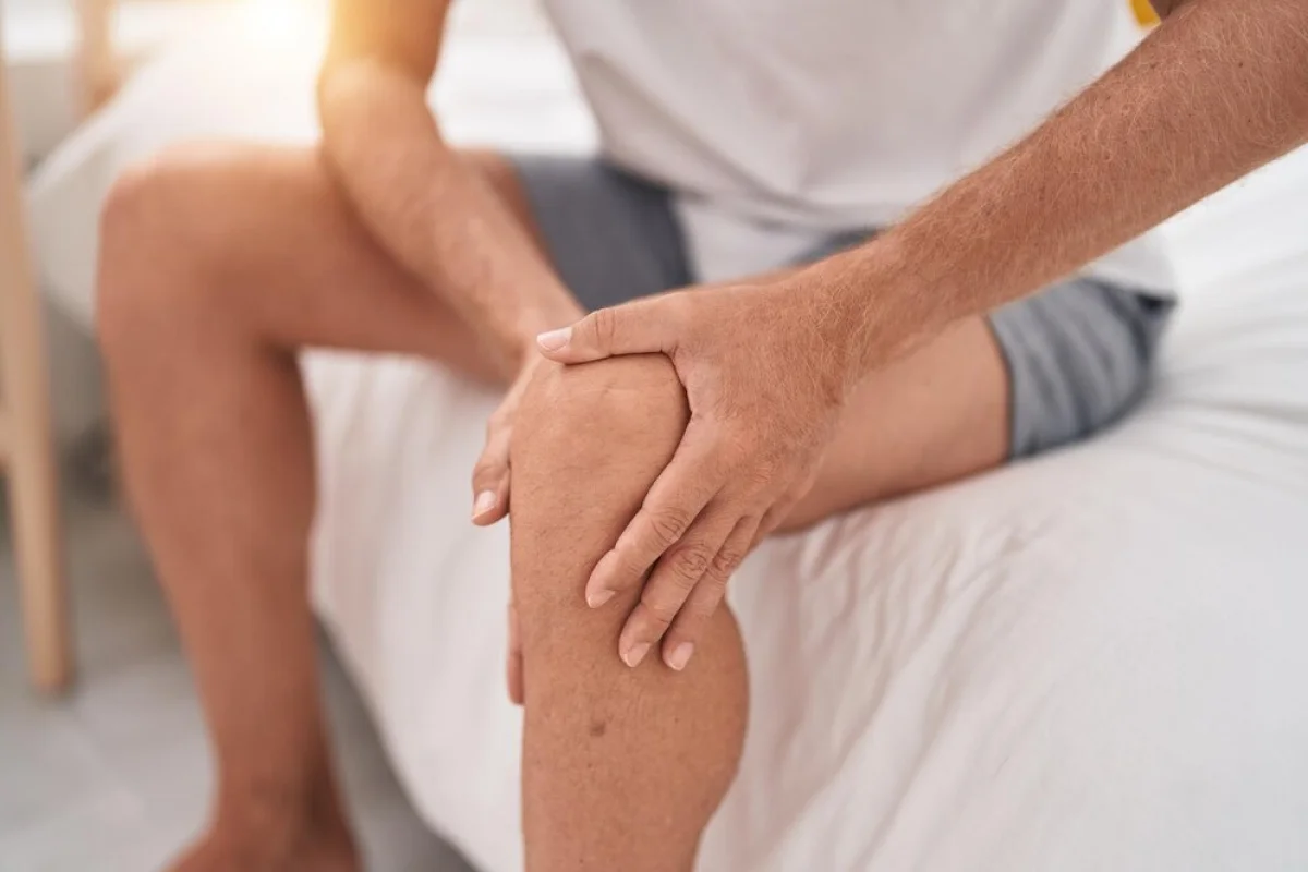 Dealing with Degenerative Arthritis 2 A man sitting on a bed, clutching his knee with both hands, indicating joint pain or discomfort.